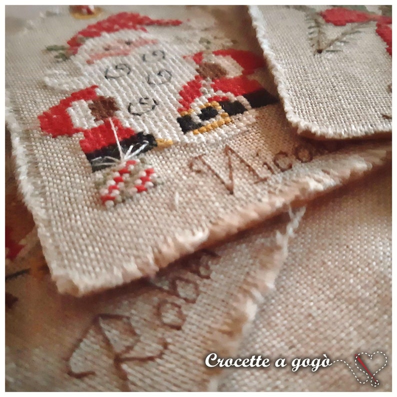Puede incluir: Primer plano de un adorno de tela beige con un dise&ntilde;o de punto de cruz de Pap&aacute; Noel sosteniendo un bast&oacute;n de caramelo. El adorno est&aacute; etiquetado como "Made" y "Crocette a gog&ograve;".