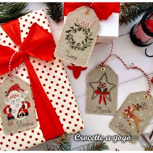 Puede incluir: Cuatro etiquetas de regalo de Navidad hechas a mano con dise&ntilde;os de punto de cruz. Las etiquetas est&aacute;n hechas de tela de lino natural y presentan una corona, una estrella, un reno y Santa Claus. Las etiquetas est&aacute;n etiquetadas con los nombres Luca, Marcola, Roberta y Nicol&aacute;s.