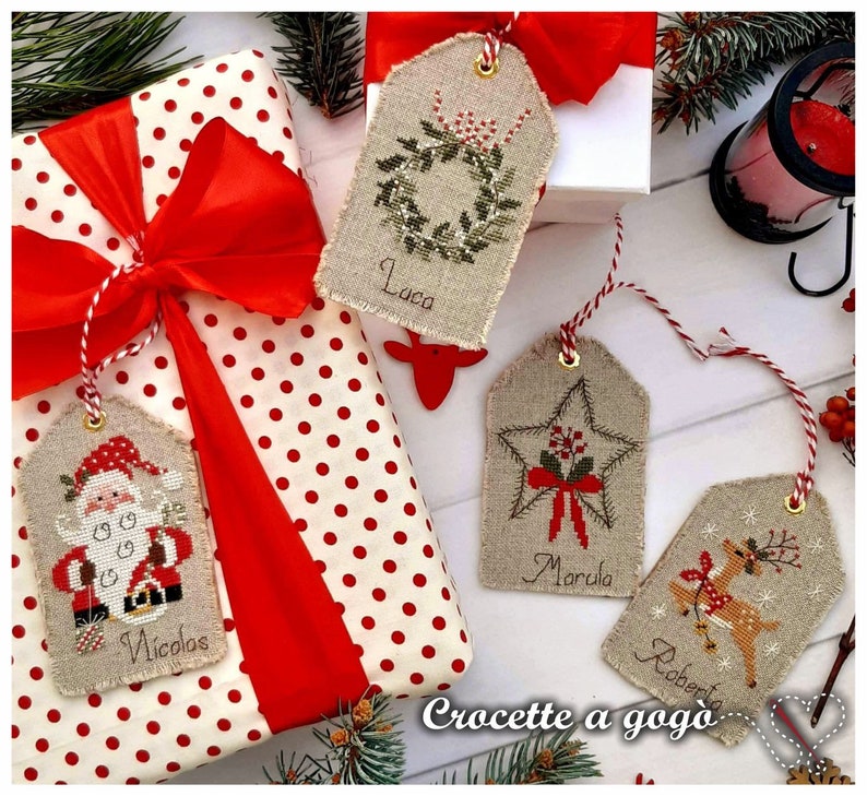 Puede incluir: Cuatro etiquetas de regalo de Navidad hechas a mano con dise&ntilde;os de punto de cruz. Las etiquetas est&aacute;n hechas de tela de lino natural y presentan una corona, una estrella, un reno y Santa Claus. Las etiquetas est&aacute;n etiquetadas con los nombres Luca, Marcola, Roberta y Nicol&aacute;s.