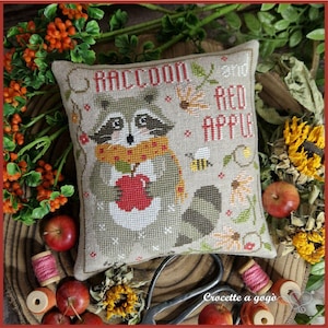 Peut inclure: Un coussin carré en lin avec une broderie au point de croix représentant un raton laveur portant une écharpe jaune et tenant une pomme rouge. Le texte indique RACCOON and RED APPLE. Le motif inclut des fleurs et une abeille sur fond beige.