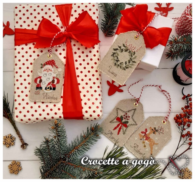 Puede incluir: Cuatro etiquetas de regalo de Navidad hechas a mano con dise&ntilde;os de punto de cruz. Las etiquetas est&aacute;n hechas de tela de lino natural y presentan un Pap&aacute; Noel, una corona, una estrella y un reno. Las etiquetas est&aacute;n atadas con un cordel rojo y blanco y tienen los nombres "Nicola", "Luca", "Maria" y "Roberto" escritos en ellas. Las etiquetas se muestran sobre un fondo blanco con una caja de regalo de lunares rojos y blancos y un peque&ntilde;o &aacute;rbol de Navidad.