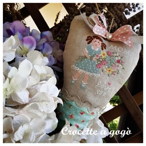 May include: A heart-shaped linen pillow with a cross-stitch design of a girl holding a bouquet of flowers. The pillow is decorated with a pink and white polka dot ribbon and a pearl. The text "Mommy I love you" is stitched on the pillow. The pillow is hanging on a wooden fence.