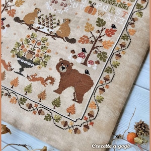 Peut inclure: Un motif de point de croix repr&eacute;sentant un ours brun, des &eacute;cureuils, des oiseaux et une vari&eacute;t&eacute; de feuillages d'automne. Le motif est brod&eacute; sur un tissu de lin beige et comprend le texte "MD 20 23".