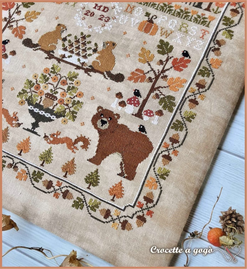 Peut inclure: Un motif de point de croix repr&eacute;sentant un ours brun, des &eacute;cureuils, des oiseaux et une vari&eacute;t&eacute; de feuillages d'automne. Le motif est brod&eacute; sur un tissu de lin beige et comprend le texte "MD 20 23".