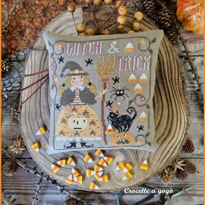 May include: A decorative, square, cross-stitched pillow with a Halloween theme. The design features a witch, a black cat, a spider, and candy corn. The words "Witch & Trick" are stitched at the top. The pillow is on a wooden surface.