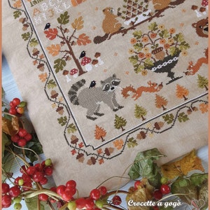 Peut inclure: Un motif de point de croix repr&eacute;sentant une sc&egrave;ne de for&ecirc;t avec un raton laveur, un &eacute;cureuil, un ours et divers arbres et champignons. Le motif est bord&eacute; d'un cadre de glands, de feuilles et d'autres &eacute;l&eacute;ments automnaux. Le texte "Crocette a gogo" est visible dans le coin inf&eacute;rieur droit.