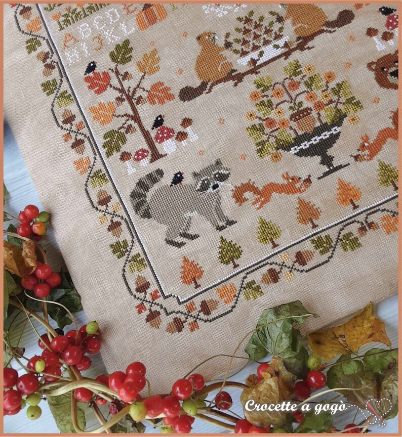 Peut inclure: Un motif de point de croix repr&eacute;sentant une sc&egrave;ne de for&ecirc;t avec un raton laveur, un &eacute;cureuil, un ours et divers arbres et champignons. Le motif est bord&eacute; d'un cadre de glands, de feuilles et d'autres &eacute;l&eacute;ments automnaux. Le texte "Crocette a gogo" est visible dans le coin inf&eacute;rieur droit.
