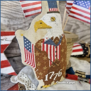 May include: A patriotic, cross-stitched eagle figurine with an American flag and the number "1776." The eagle is brown, white, and beige, with a yellow beak and a hat in red, white, and blue. The text "Crocette a gog&ograve;" is visible.