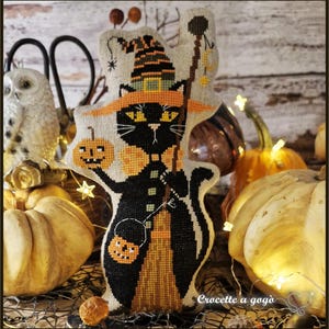 May include: A cross-stitched fabric ornament featuring a black cat wearing a striped witch hat and orange collar. The cat holds a small pumpkin in one paw and a broomstick in the other, with a jack-o'-lantern bucket hanging from its arm.