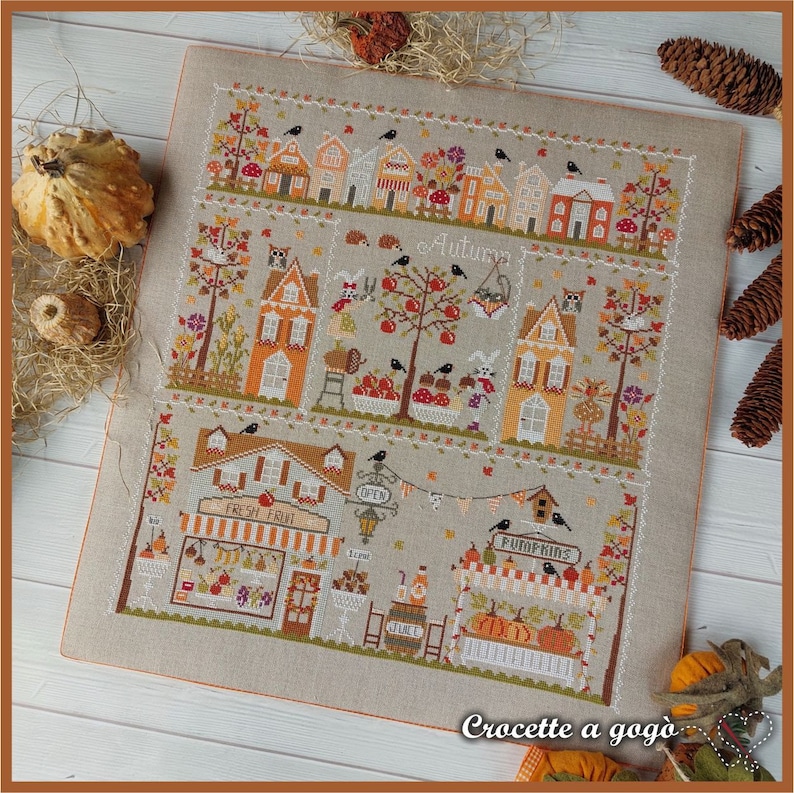 May include: A cross-stitch pattern featuring a charming autumn village scene with houses, trees, pumpkins, and a sign that reads "Crochette a gogo".