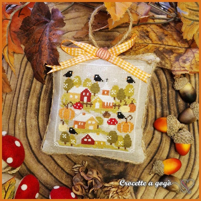 Op de afbeelding: Een vierkant, kruissteek ornament met een dorpssc&egrave;ne met rode huizen, pompoenen en bomen. Het ornament is afgewerkt met een stof die op jute lijkt en heeft een ruitjeslint en een jute lus om op te hangen. Eikels en paddenstoelen omringen het ornament.