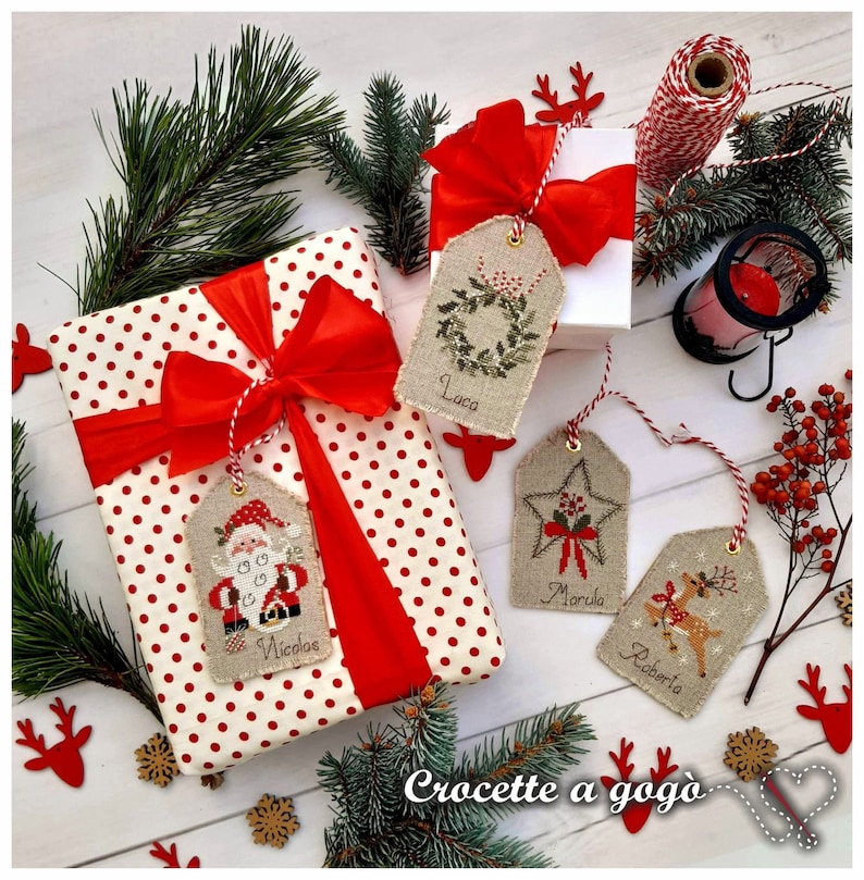 Puede incluir: Tres etiquetas de regalo de Navidad con hilo rojo y blanco, que presentan dise&ntilde;os bordados de Pap&aacute; Noel, un reno y una corona. Las etiquetas est&aacute;n etiquetadas como "Nicola", "Roberta" y "Luca". Las etiquetas est&aacute;n atadas a una caja de regalo de lunares rojos y blancos con una cinta roja.