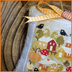 Op de afbeelding: Een kruissteekornament met herfstthema, met een dorpsgezicht met pompoenen, bomen en huizen. Het ontwerp omvat een rood huis, zwarte vogels en de tekst "Crocette a gog&ograve;". Het is versierd met een ruitjeslint en een pompoenvormige versiering.