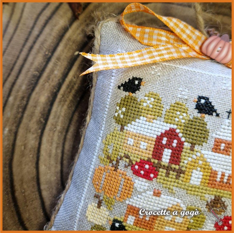 Op de afbeelding: Een kruissteekornament met herfstthema, met een dorpsgezicht met pompoenen, bomen en huizen. Het ontwerp omvat een rood huis, zwarte vogels en de tekst "Crocette a gog&ograve;". Het is versierd met een ruitjeslint en een pompoenvormige versiering.