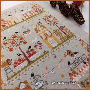 May include: A cross-stitch pattern featuring a charming autumn scene with houses, trees, and birds. The text "Croquette a gogo" is visible in the bottom right corner.