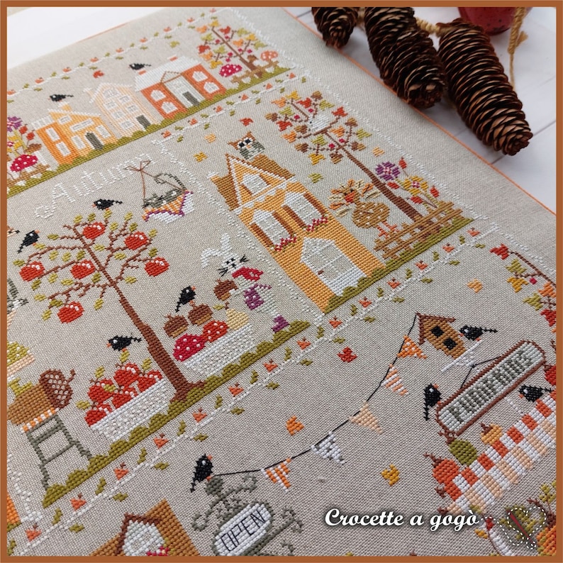 May include: A cross-stitch pattern featuring a charming autumn scene with houses, trees, and birds. The text "Croquette a gogo" is visible in the bottom right corner.