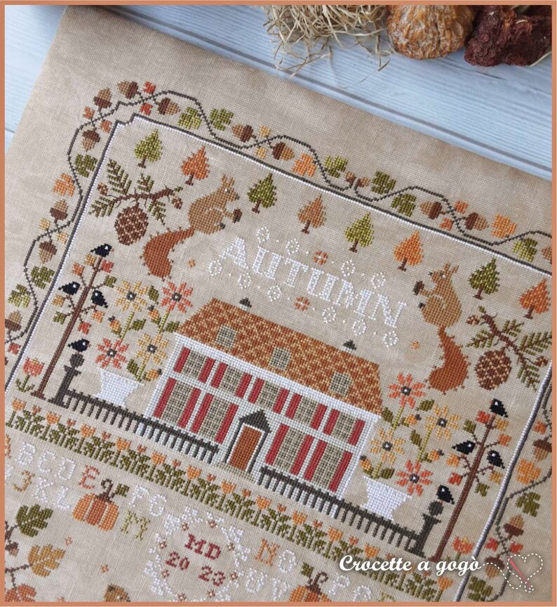 Peut inclure: Mod&egrave;le de point de croix repr&eacute;sentant une charmante sc&egrave;ne d'automne avec une maison, des arbres, des &eacute;cureuils, des oiseaux et des citrouilles. Le motif est intitul&eacute; "Automne" et comprend le texte "Crocette a gogo".