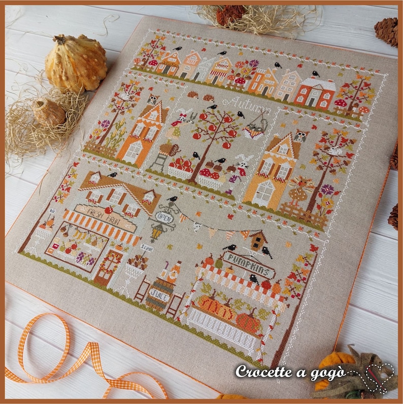 May include: Cross-stitch pattern featuring a charming autumn village scene with houses, trees, birds, and a pumpkin patch. The pattern includes the text "Autumn" and "Crocquette a gogo".