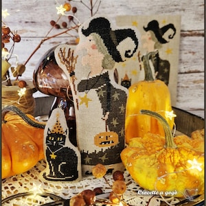 May include: A cross-stitch embroidery of a witch wearing a black hat and holding a broom, alongside a black cat wearing a yellow hat. The witch wears a dark dress with star patterns and holds a small orange pumpkin. The design is on light fabric.
