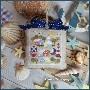 Può includere: Un ornamento quadrato in lino con una scena costiera a punto croce raffigurante case, un faro, una barca a vela e un granchio. Il pezzo è sormontato da un fiocco in nastro blu scuro e un ciondolo a forma di barca a vela, appeso a un cordino.
