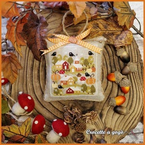 Può includere: Un ornamento a cuscino ricamato a punto croce raffigurante una scena di villaggio autunnale con case rosse, zucche e alberi. Il cuscino è decorato con un nastro a quadretti e un anello di iuta. Ghiande decorative e funghi di feltro circondano il cuscino.