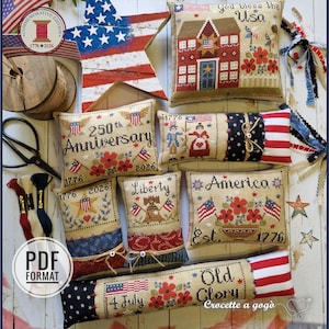 May include: A selection of cross-stitch cushions and decorative pieces with a patriotic theme. The cushions display American flags, floral patterns, and phrases like "250th Anniversary" and "Liberty." The colour scheme is red, white, and blue, with a rustic appearance.