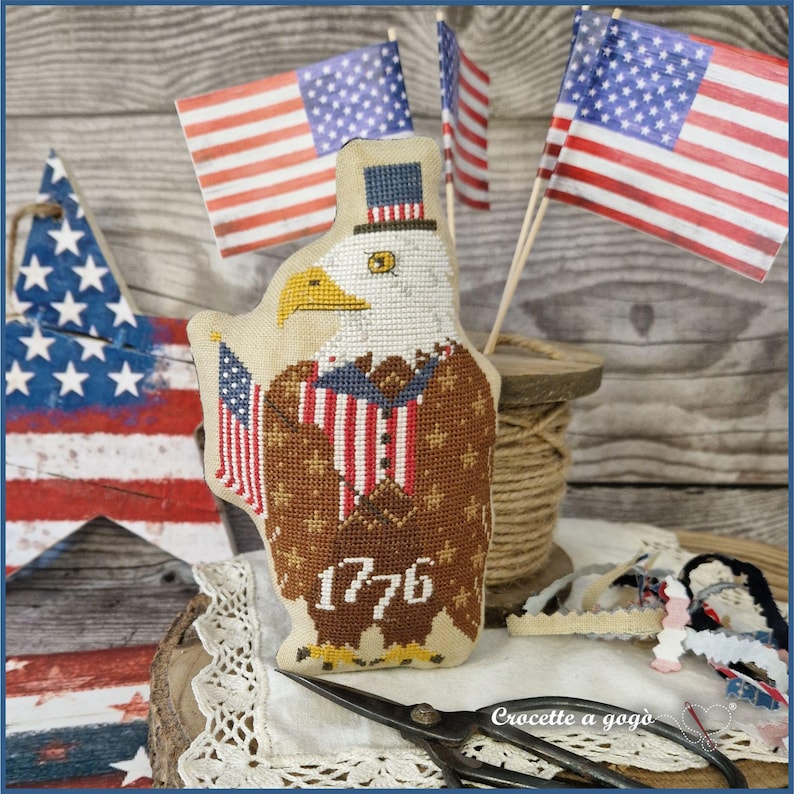 May include: A patriotic, handcrafted eagle-shaped ornament. The eagle is adorned with a top hat and a suit featuring red, white, and blue stripes, holding a small American flag. The number "1776" is stitched on the eagle's chest. Miniature American flags are in the background.