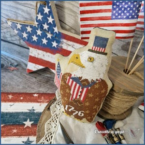 May include: A patriotic-themed display featuring a wooden star with a painted American flag design, a stitched eagle holding flags, and miniature American flags. The eagle has a top hat and the year "1776" stitched on its chest. The color palette includes red, white, and blue.