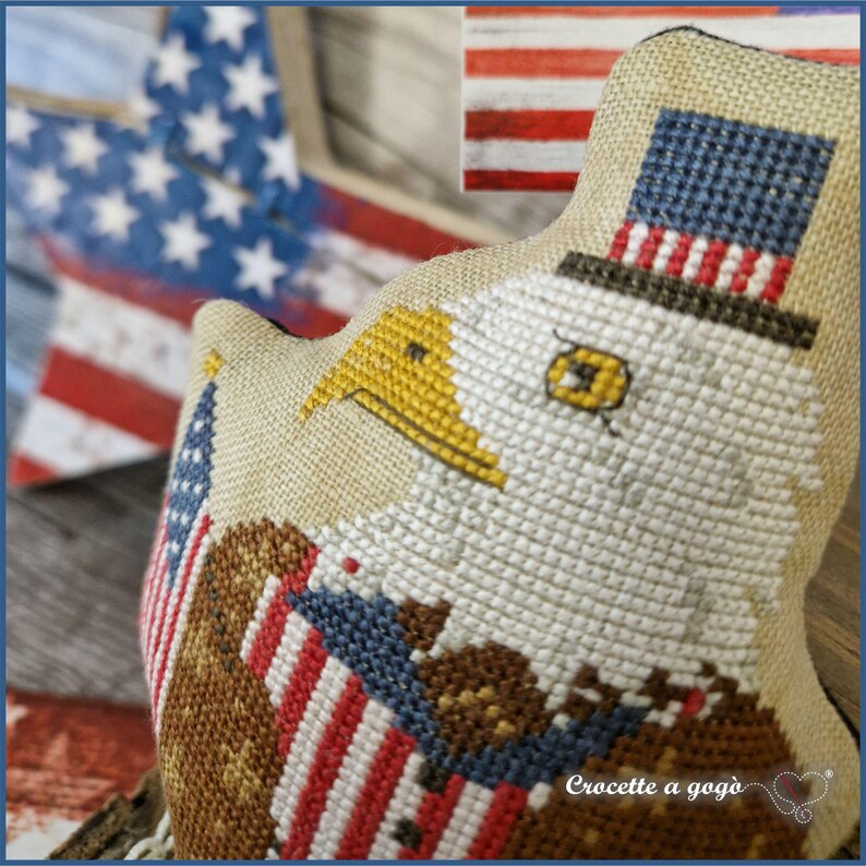 May include: A patriotic, cross-stitched eagle wearing a top hat with a US flag design. The eagle has a yellow beak and is holding a flag. The background includes a wooden star and a flag, both with a distressed look.