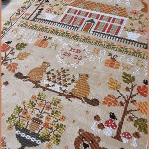 Peut inclure: Mod&egrave;le de point de croix repr&eacute;sentant une charmante sc&egrave;ne d'automne avec une maison, des arbres, des &eacute;cureuils et un ours. Le motif comprend le texte "Autumn" et "Crocette a gogo".