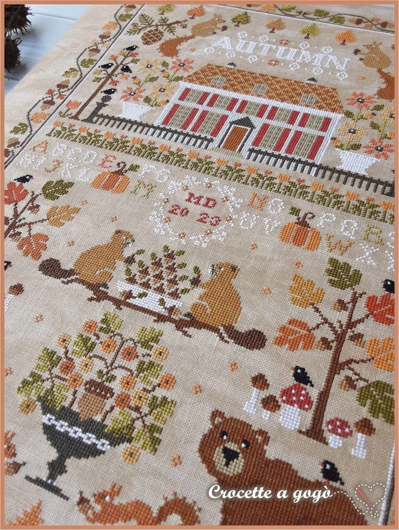 Peut inclure: Mod&egrave;le de point de croix repr&eacute;sentant une charmante sc&egrave;ne d'automne avec une maison, des arbres, des &eacute;cureuils et un ours. Le motif comprend le texte "Autumn" et "Crocette a gogo".