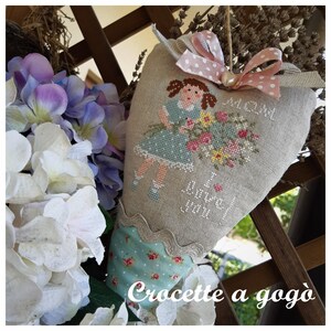 May include: A heart-shaped pillow with a cross-stitch design of a girl holding a bouquet of flowers. The pillow is made of linen and has a scalloped edge. The text "I love you" is stitched on the pillow. The pillow is decorated with a pink ribbon and a pearl.