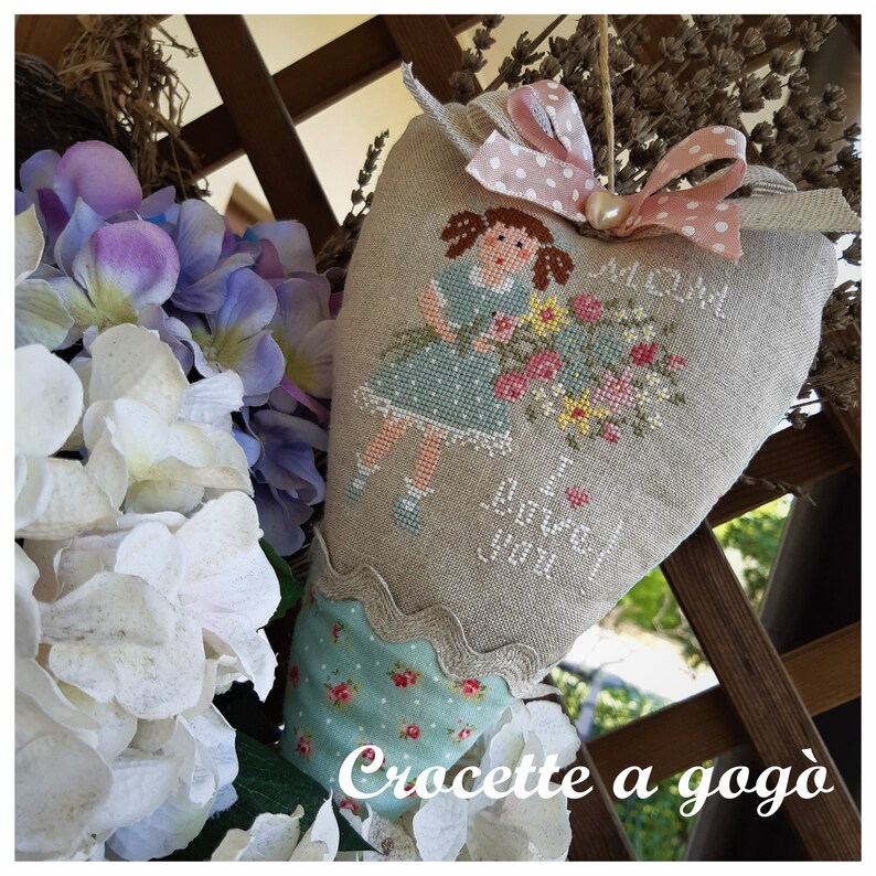 May include: A heart-shaped pillow with a cross-stitch design of a girl holding a bouquet of flowers. The pillow is made of linen and has a scalloped edge. The text "I love you" is stitched on the pillow. The pillow is decorated with a pink ribbon and a pearl.