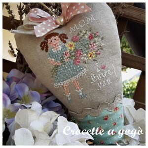 May include: A heart-shaped linen pillow with a cross-stitch design featuring a girl holding a bouquet of flowers. The pillow is decorated with a pink and white polka dot ribbon and a pearl. The text "Mom I love you" is stitched on the pillow. The pillow is sitting on a table with a white crocheted doily.