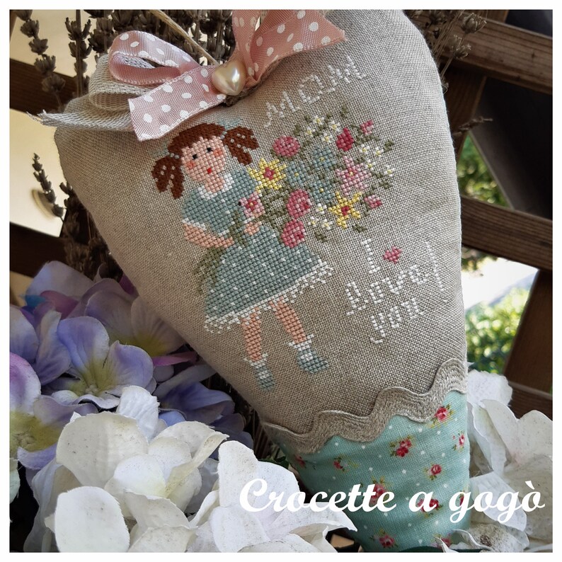 May include: A heart-shaped linen pillow with a cross-stitch design featuring a girl holding a bouquet of flowers. The pillow is decorated with a pink and white polka dot ribbon and a pearl. The text "Mom I love you" is stitched on the pillow. The pillow is sitting on a table with a white crocheted doily.