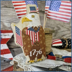 May include: A patriotic, cross-stitched eagle with an American flag design. The eagle wears a top hat and has the year "1776" stitched on its chest. The background includes American flags and sewing supplies, creating a handcrafted theme.