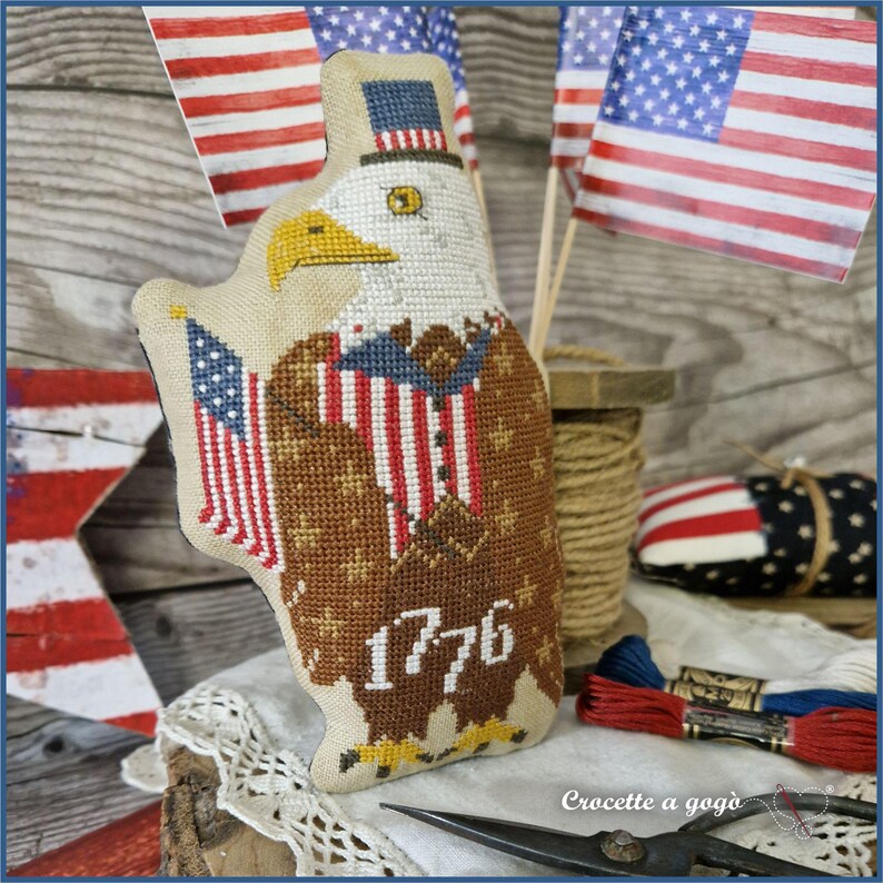 May include: A patriotic, cross-stitched eagle with an American flag design. The eagle wears a top hat and has the year "1776" stitched on its chest. The background includes American flags and sewing supplies, creating a handcrafted theme.