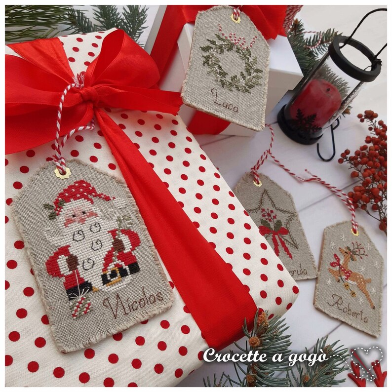Puede incluir: Una caja de regalo blanca con lunares rojos y una cinta de sat&eacute;n roja. La caja est&aacute; decorada con cuatro etiquetas de regalo de Navidad hechas a mano con dise&ntilde;os de punto de cruz. Las etiquetas est&aacute;n etiquetadas "Laca", "Nicolas", "Roberta" y "Annbia". El texto "Crocette a gog&ograve;" est&aacute; escrito en la caja.