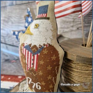 May include: A cross-stitched patriotic eagle ornament. The eagle is wearing a top hat and a suit with the American flag design. The ornament is brown, white, red, and blue, with the number "1776" stitched on the bottom.