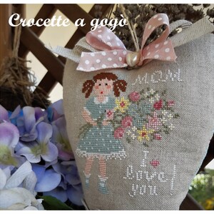 May include: A heart-shaped linen pillow with a cross-stitch design of a girl holding a bouquet of flowers. The pillow is decorated with a pink ribbon with white polka dots and the text "I love you" is stitched on the bottom.