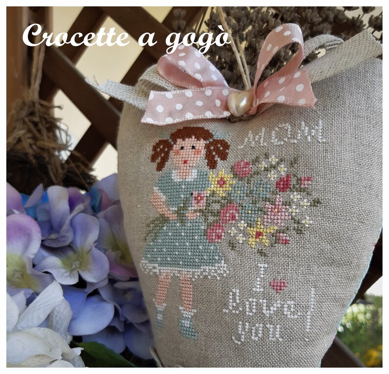 May include: A heart-shaped linen pillow with a cross-stitch design of a girl holding a bouquet of flowers. The pillow is decorated with a pink ribbon with white polka dots and the text "I love you" is stitched on the bottom.