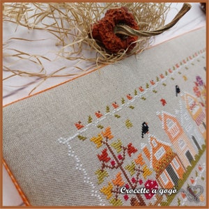 May include: Cross-stitch pattern of a fall scene with houses, trees, and birds. The pattern is stitched on a beige linen fabric with orange stitching around the edges. The text "Crocette a gogo" is stitched in the bottom right corner.