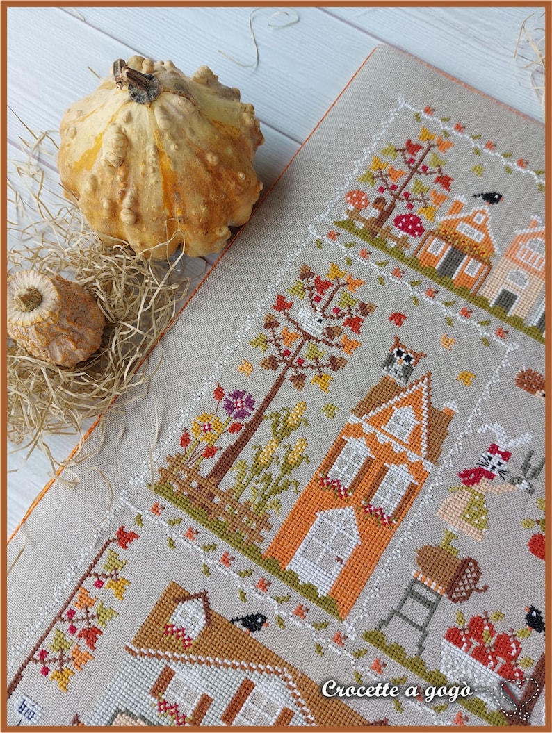 May include: A cross-stitch pattern featuring three houses with orange and brown accents, surrounded by trees, flowers, and a bird. The text "Crochette a gogo" is visible in the bottom right corner.
