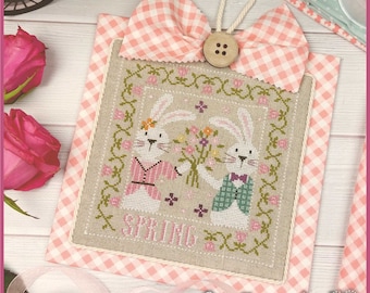 May include: A cross-stitch embroidery design featuring two bunnies holding a bouquet of flowers. The design is framed with a pink and white gingham border and the word "SPRING" is stitched in the center. The design is titled "Crocette a gogo".