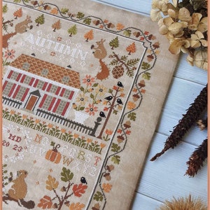 Peut inclure: Un sampler de point de croix beige avec une maison brune, des arbres et des animaux. Le texte "AUTUMN BLESSINGS" est brod&eacute; en fil blanc.