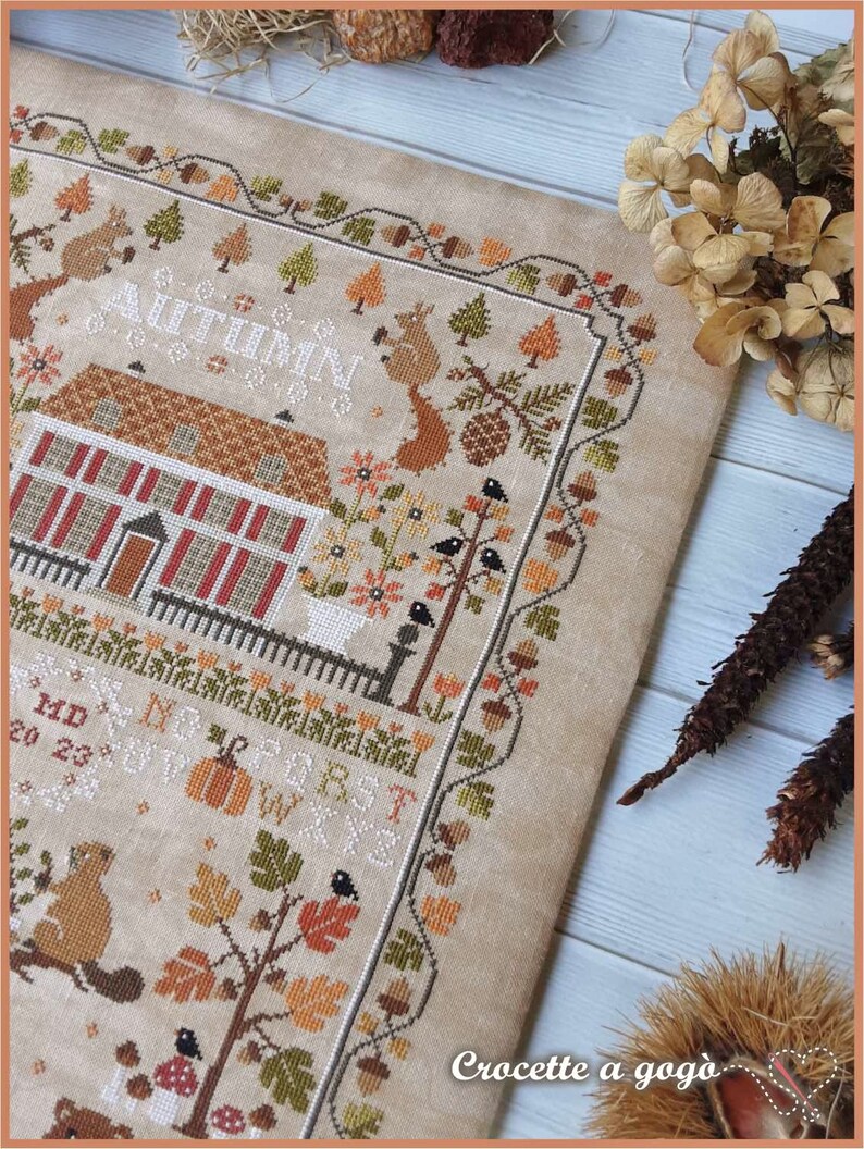 Peut inclure: Un sampler de point de croix beige avec une maison brune, des arbres et des animaux. Le texte "AUTUMN BLESSINGS" est brod&eacute; en fil blanc.