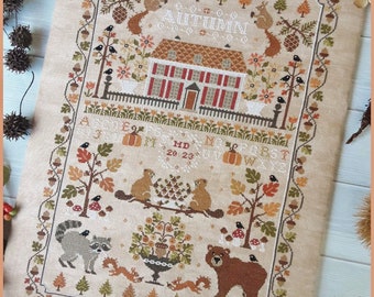 May include: Cross-stitch pattern featuring a house with a red roof and a garden with a bear, raccoons, squirrels, and birds. The pattern is titled "Autumn" and includes the letters of the alphabet and the numbers 20-23. The pattern is on a beige fabric.