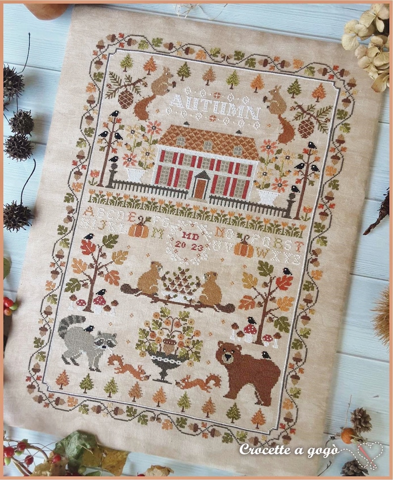 Peut inclure: Mod&egrave;le de point de croix repr&eacute;sentant une maison avec un toit rouge et un jardin avec un ours, des ratons laveurs, des &eacute;cureuils et des oiseaux. Le motif est intitul&eacute; "Automne" et comprend les lettres de l'alphabet et les chiffres 20 &agrave; 23. Le motif est sur un tissu beige.