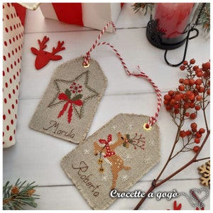 Puede incluir: Dos etiquetas de regalo de Navidad hechas a mano con cuerda roja y blanca. La etiqueta de la izquierda tiene una estrella roja con un lazo verde y el nombre "Marula" escrito debajo. La etiqueta de la derecha tiene un reno rojo con una corona verde y el nombre "Roberta" escrito debajo. Las etiquetas est&aacute;n sobre una superficie de madera blanca con un fondo de lunares rojos y blancos.