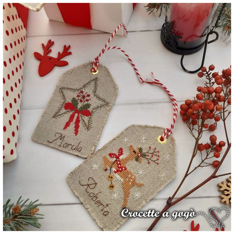 Puede incluir: Dos etiquetas de regalo de Navidad hechas a mano con cuerda roja y blanca. La etiqueta de la izquierda tiene una estrella roja con un lazo verde y el nombre "Marula" escrito debajo. La etiqueta de la derecha tiene un reno rojo con una corona verde y el nombre "Roberta" escrito debajo. Las etiquetas est&aacute;n sobre una superficie de madera blanca con un fondo de lunares rojos y blancos.